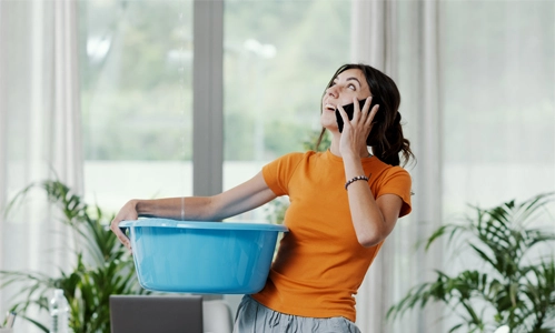 woman on the phone holding bucket catching dripping water from ceiling